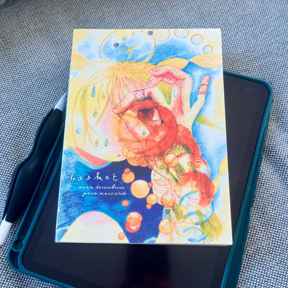 Basket, a graphic novel by marie derambure and paco moccand. The cover is a textured card stock printed with sketchy art in vibrant yellows, oranges and blues. A cartoon girl with blonde hair and brown eyes peers at the viewer through a hole she’s made with her pointer finger and thumb. The book lays on a digital tablet in a green case next to an Apple Pencil. The pile sits on a gray and white, textured upholstery fabric.