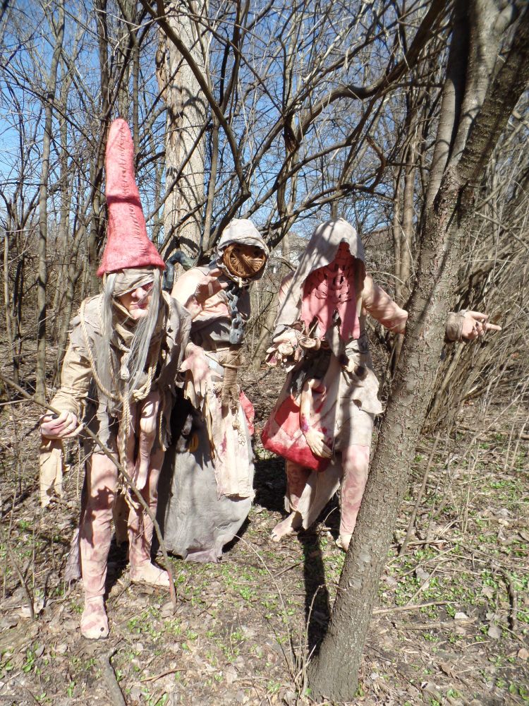 A brown, leafless wooded area with three figures cast in shadows from the bare branches. Weavess, Brewess, and Whispess, the Crones of Crookback Bog from Witcher 3, lean forward menacingly in their ragged brown and red clothes. Weavess wears a tall, conical red hat and badages across her face. Her large nose and chin protruding, while tiny legs hang from her belly. Brewess, the tallest, stares forward with her basket mask obscuring her face. Whispess gestures, her bag stained red with blood, an arm protruding, with her woven red mask also hiding her face.