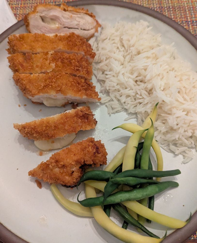 Breaded and sliced chicken cutlet oozing cheese on a white plate with some rice and string beans on the side