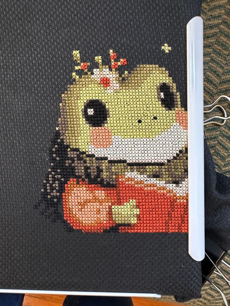 Image is of a frog reading a rust-colored book, counted cross stitch on black cloth. Frog is wearing alike a rust-colored sweater and a black and khaki-green scarf. 
There’s a sort of little flower behind his head at the top. 