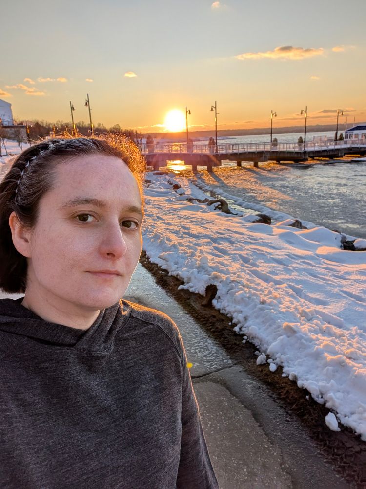 Selfie of me in front of the icy Potomac River with the setting sun behind me.  There is some sort of boardwalk that is going into the river and snow on the ground.