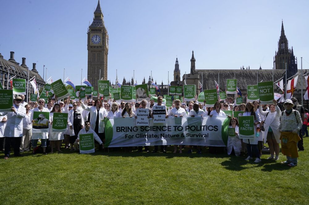 Around 200 scientists in front of the UK Houses of Parliament. Scientists are wearing white lab coats, assembled behind a banner reading "Bring Science to Politics... For climate, nature and our survival" 