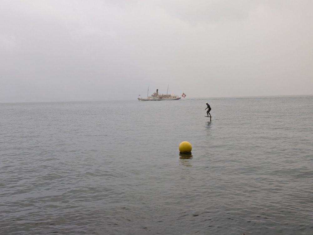 Tourist ship in the background, man on a foilboard, yellow buoy in foreground on a grey, rainy day, lake Geneva.
