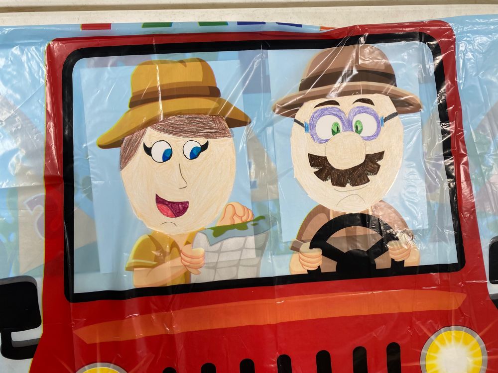 This image features a colorful cartoon illustration of two characters sitting in the front seats of a red vehicle, possibly a jeep. The scene is viewed from the outside, looking through the windshield. The character on the left, who appears to be a woman, has light skin, bright blue eyes, and short brown hair peeking out from under a yellow safari-style hat. She is smiling enthusiastically while holding and examining a map, suggesting she might be navigating. She wears a yellow shirt and seems excited about the journey.

The character on the right is a man with light skin, thick brown eyebrows, green eyes, and a large round nose. He sports a thick brown mustache and wears purple-rimmed glasses along with a brown safari hat matching his tan shirt. He has a serious, somewhat concerned expression, both hands gripping the steering wheel, focused on driving. The background shows a bright, clear sky, adding to the adventure theme, with vivid colors and playful design.