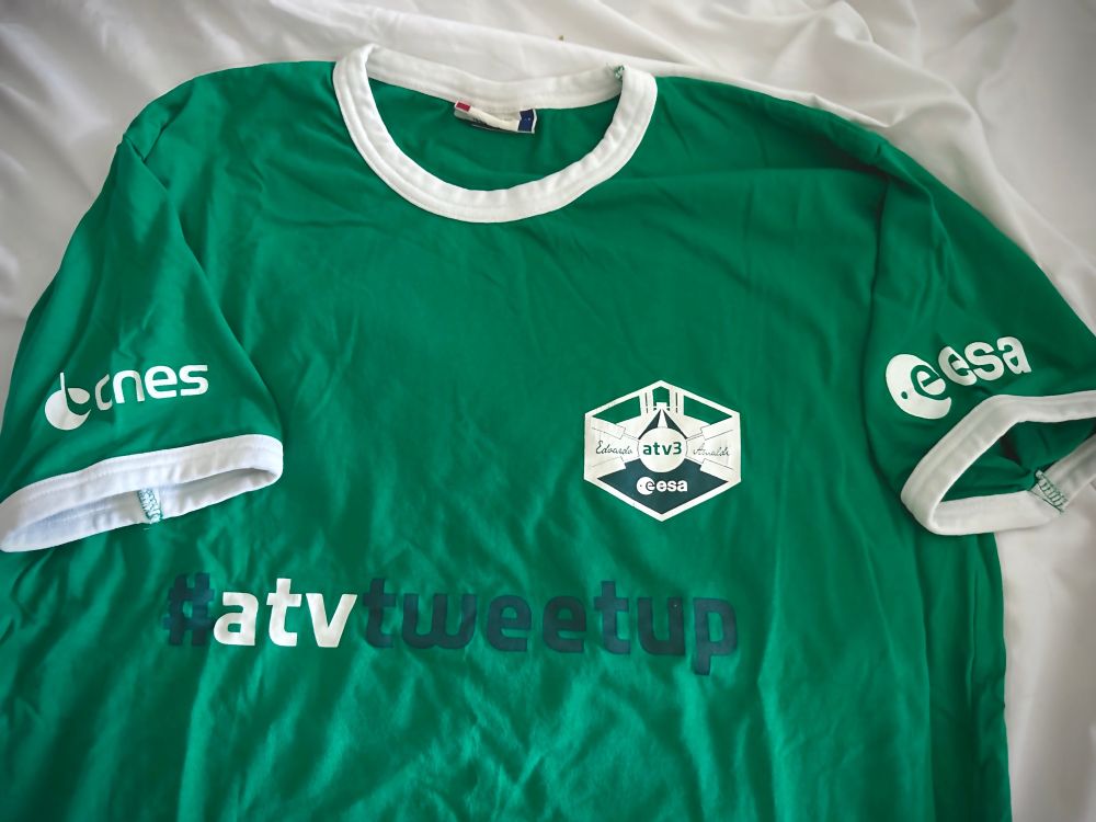 Photo of a green ringer tshirt with CNES logo on the left sleeve, ESA logo on the right sleeve. 

The mission logo has a stylized view of the ISS and Edoardo (atv3) Amaldi
#atvtweetup across the chest. 
