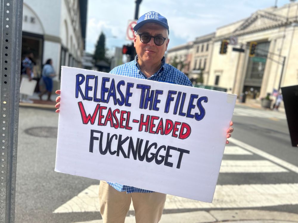Handsome man in a blue shirt and cap with a rakish smirk holds a sign: “Release the files weasel-headed fucknugget.”