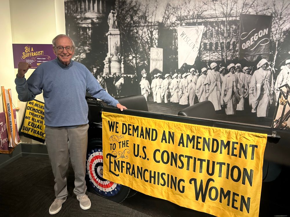 Adorable older man in blue pullover with raised fist next to a 1920s car with a yellow banner: “We demand an amendment to the U.S. Constitution enfranchising women.”