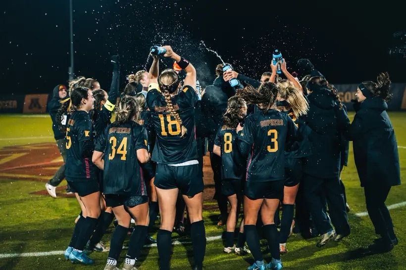 Group picture of the Gopher soccer team in their all black uniforms with gold numbers/lettering. They’re all celebrating in a circle and throwing water in the air after a big win at Elizabeth Lyle stadium at night