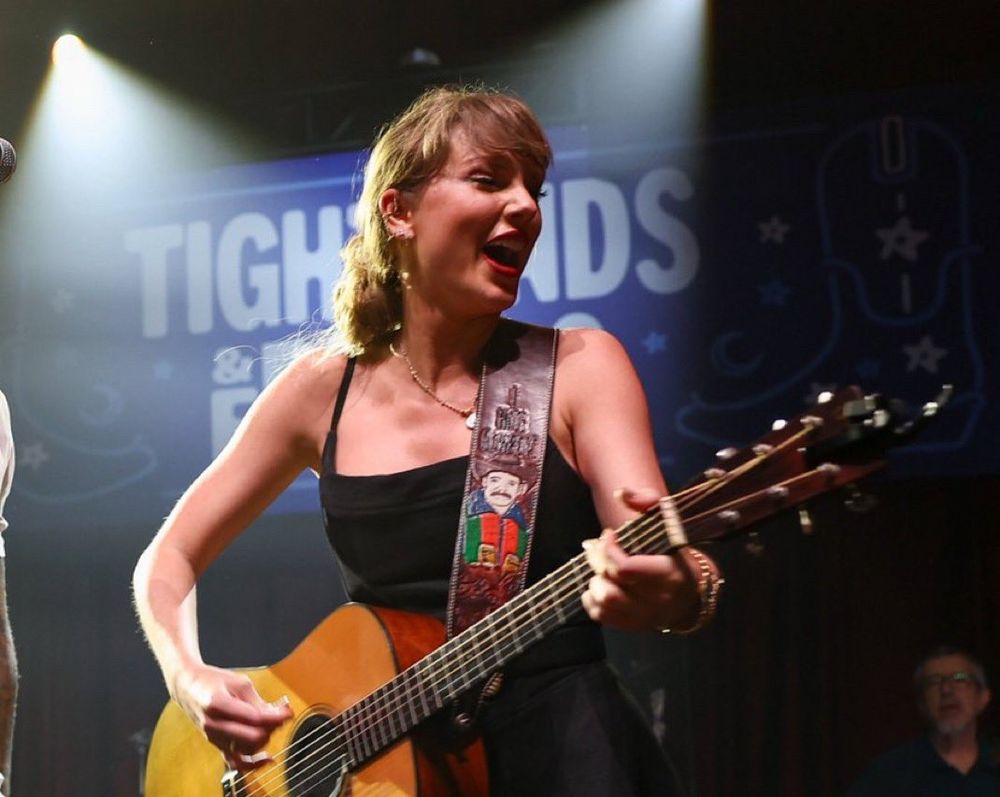 Taylor Swift from her latest performance in a black spaghetti strap dress. She is playing a light brown guitar and on stage smiling with her mouth open, looking downward 