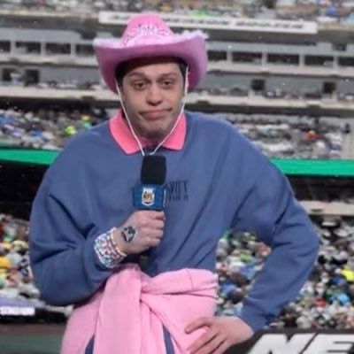 Pete davidson in a pink cowboy hat, taylor swift crew neck, and friendship bracelets holding a mic with his hand on his hip looking disappointed 