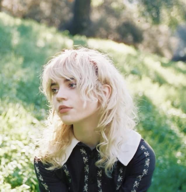 Sophie Thatcher with blonde hair, medium length with messy bangs in a black long sleeve with a white wide collar. She is sitting in the grass staring longingly to the side with her mouth slightly open