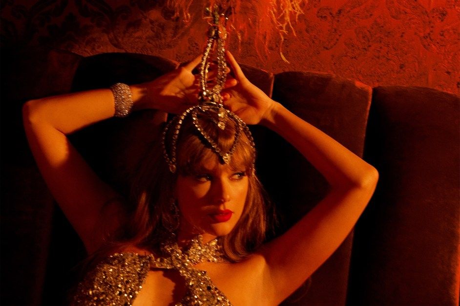 Taylor Swift with a showgirl headpiece on and a bedazzled crisscrossed top, she has her hands above her head looking away from the camera off in the distance with a straight face with red lighting 
