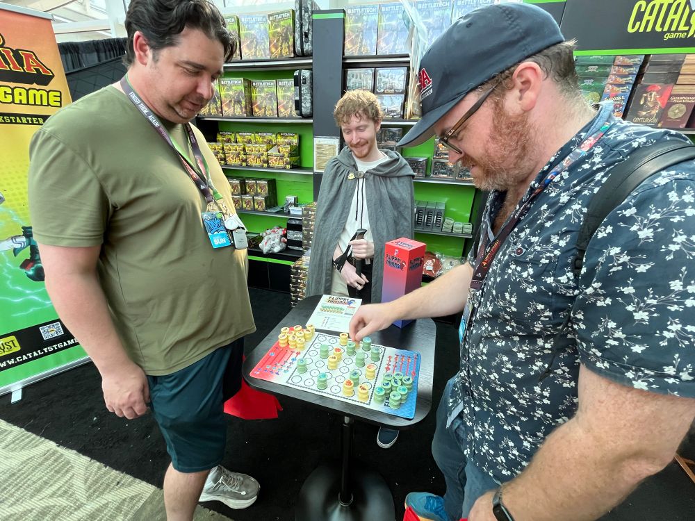 Zoomed out photo of two guys playing the game, with a third one in a cloak observing