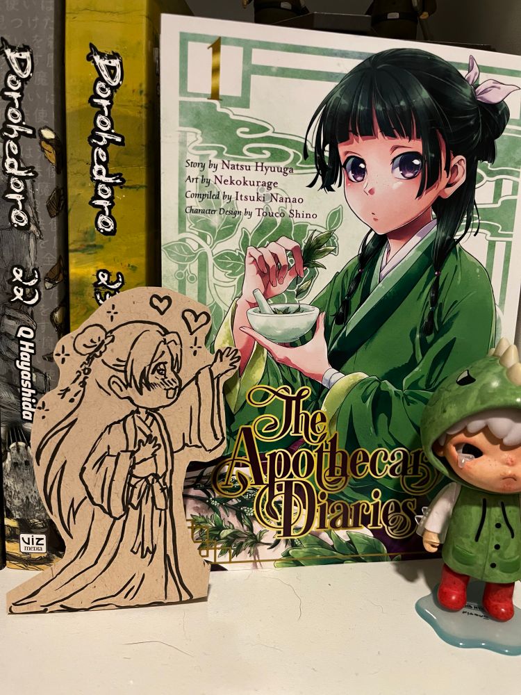 Photo of vol 1 of the Apothecary Diaries manga displayed frontward on a bookshelf. Cut out of brown sketchbook paper is a little ink drawing of chibi Jinshi staring and reaching out adoringly to the MaoMao cover illustration
