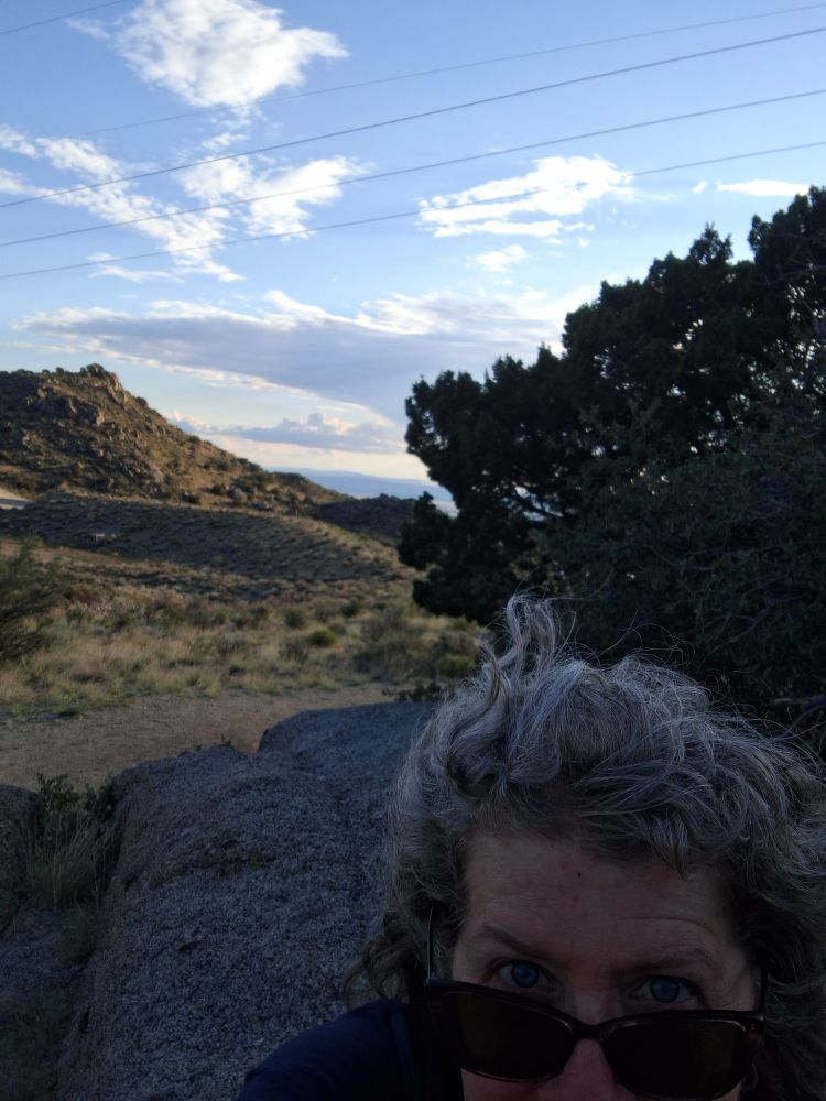 In the shaded foreground, a white woman peeks over sunglasses, her hair blows in the wind. Sandia foothills in the background with a bright blue sky and clouds above.