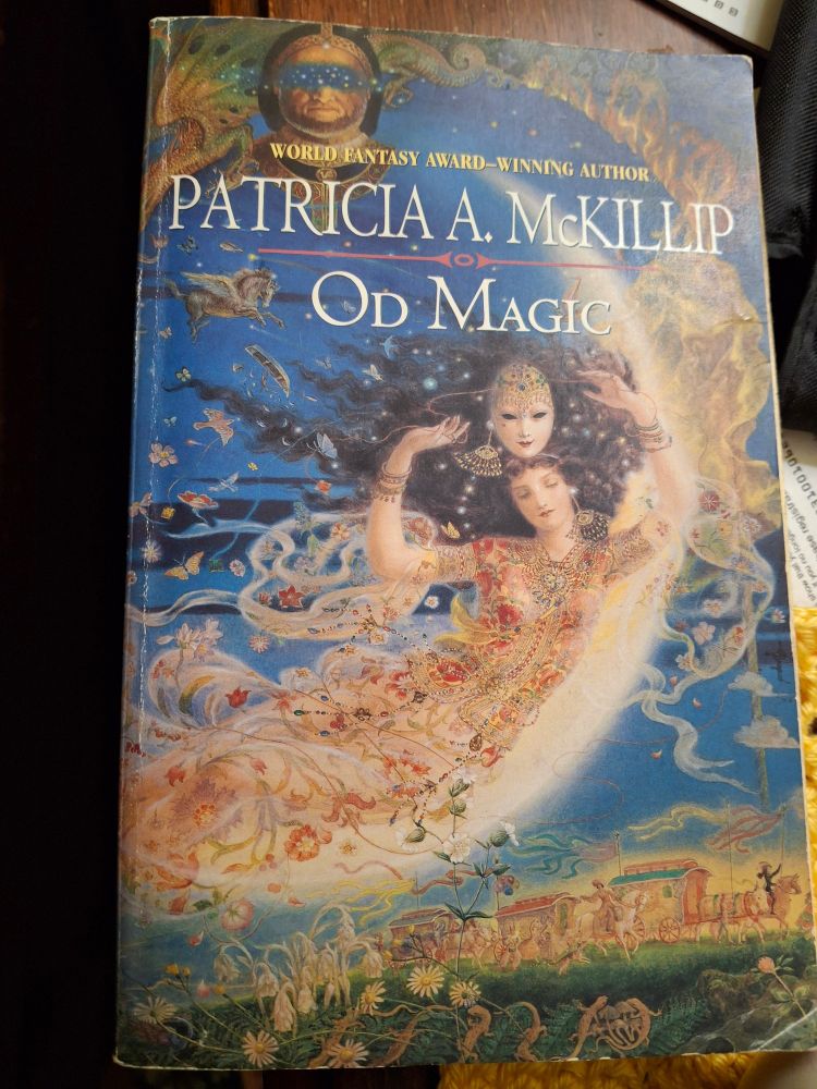 Front cover of Patricia McKillip's 2005 novel Od Magic. A young woman floats in the curve of the crescent moon, holding a mask above her head. A face with a blindfold of stars floats above and a caravan appears below.