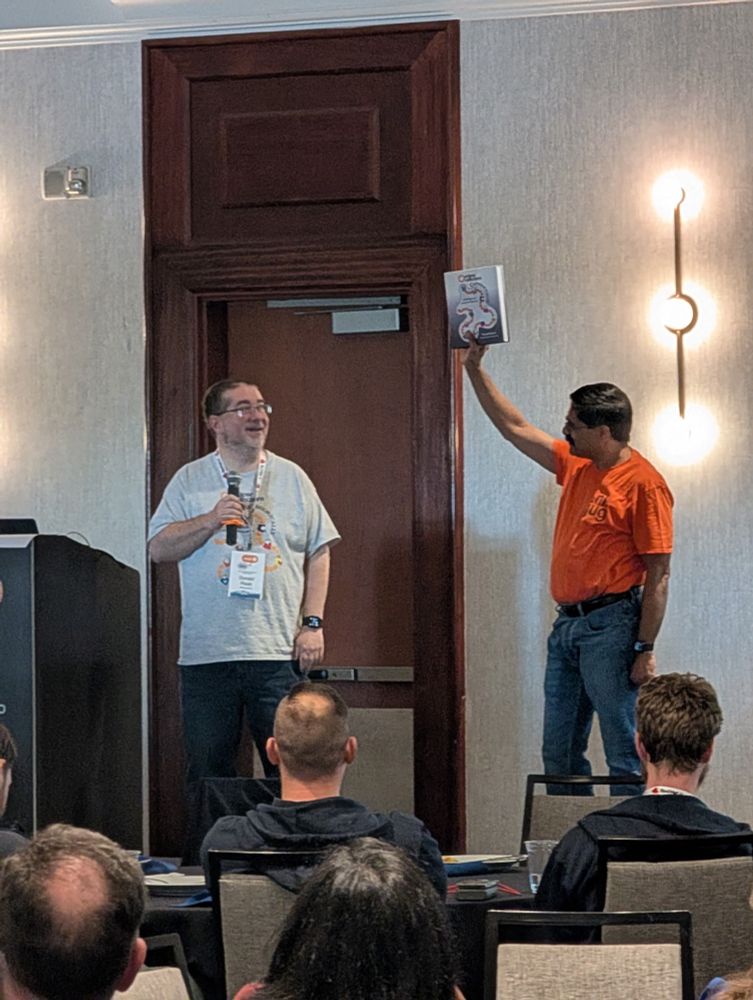 Me holding a microphone on stage telling the story of writing and publishing my first book while Venkat holds a hardcover copy of the book  up in the air about to give away to an audience member.
