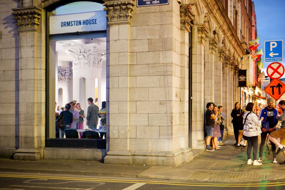 The exterior of Ormston House at twilight. A crowd of people talk to one another outside. Through a window to the left, more people can be seen.