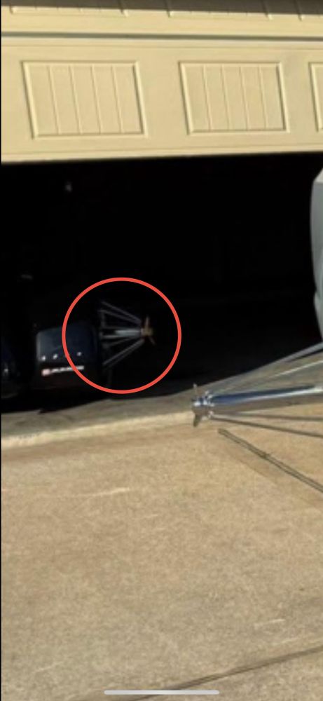 A suburban driveway. A Cadillac EV is parked in it with ridiculous chrome rims with long center shafts that stick out more than a foot from each wheel. Each shaft is supported by chrome spokes. In the background the garage interior is visible. There is another vehicle, possibly a black trike, with similar rims.