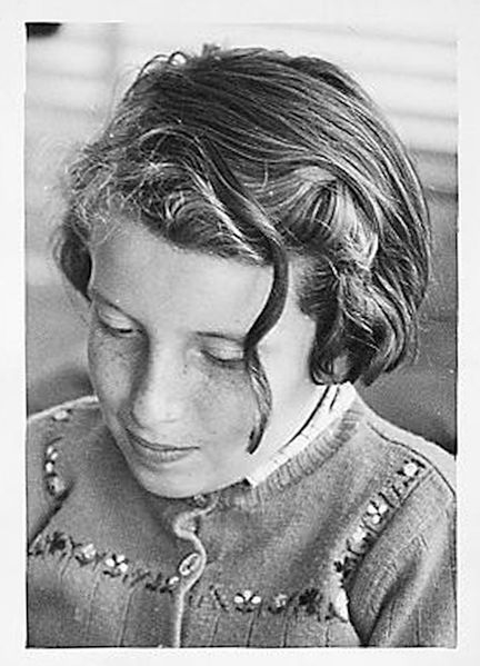 Black and white picture of my mother when she was a little girl. She's looking down and she wores a knitted cardigan with little flowers. She has short hair and a curl in front of her face. And freckles. She's smiling.