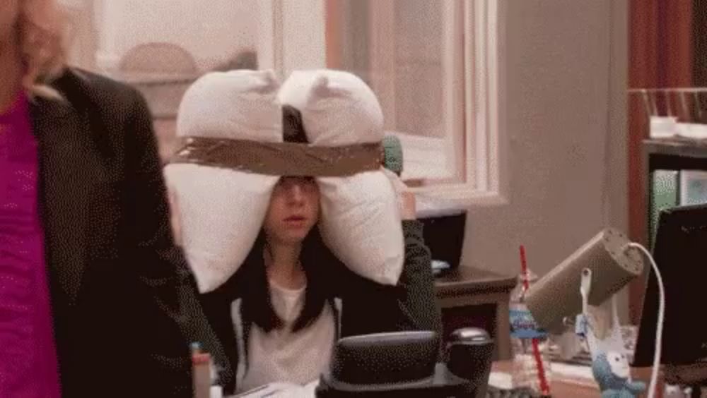 a woman is sitting at a desk with a pillow on her head and a woman is standing behind her .