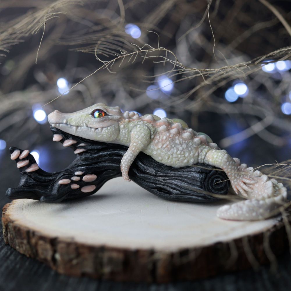 White alligator sculpture laying on a black log with mushrooms. Twinkly lights and twigs in the background 