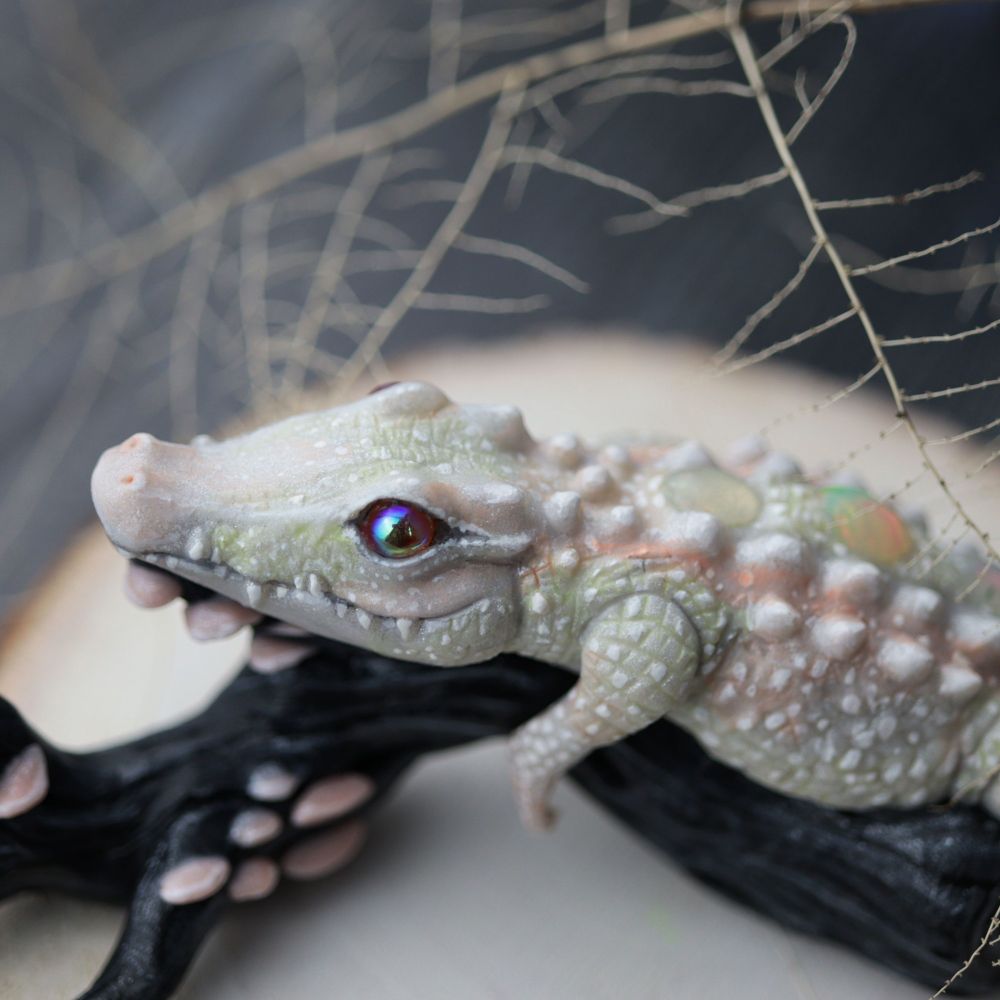 Top view. White alligator sculpture laying on a black log with mushrooms. Twigs above