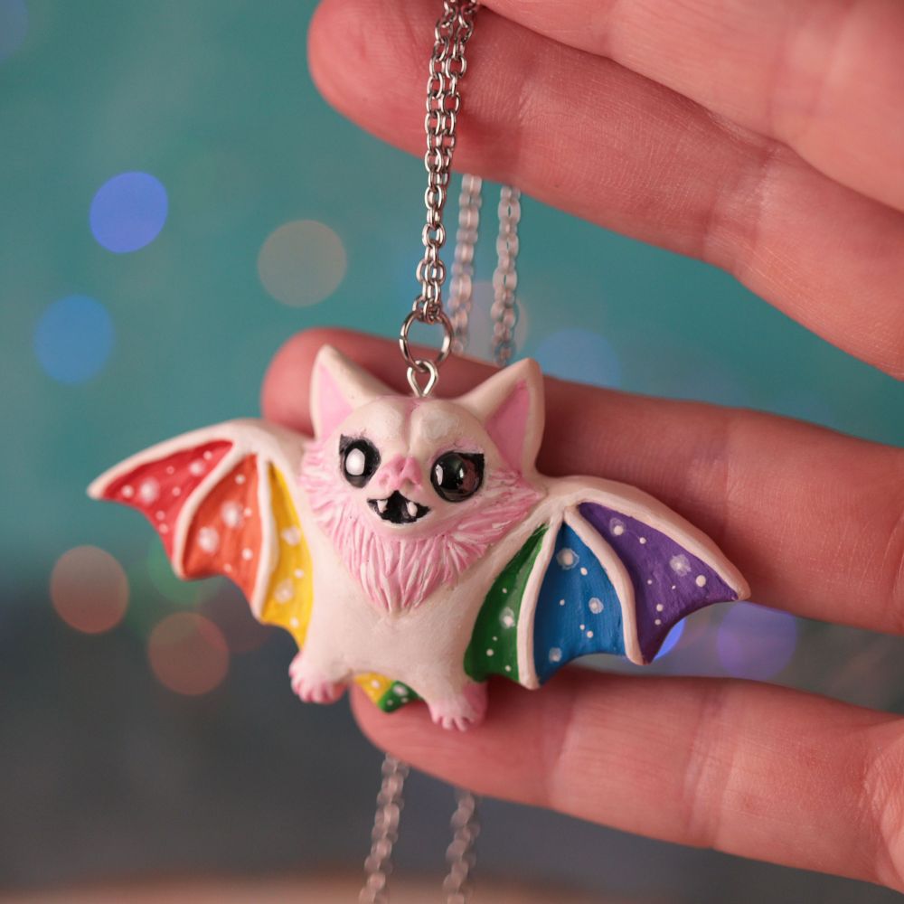 A hand holding a rainbow colored bat pendant on a chain