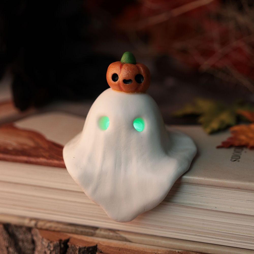 A tiny ghost with green light in its eyes and a happy pumpkin on its head. It sits on a book patiently waiting for its new haunt 