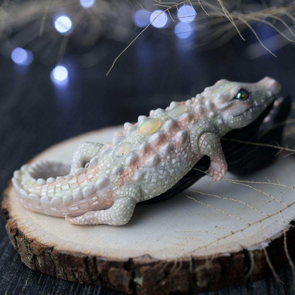 Backside. White alligator sculpture laying on a black log with mushrooms. Twinkly lights and twigs in the background 