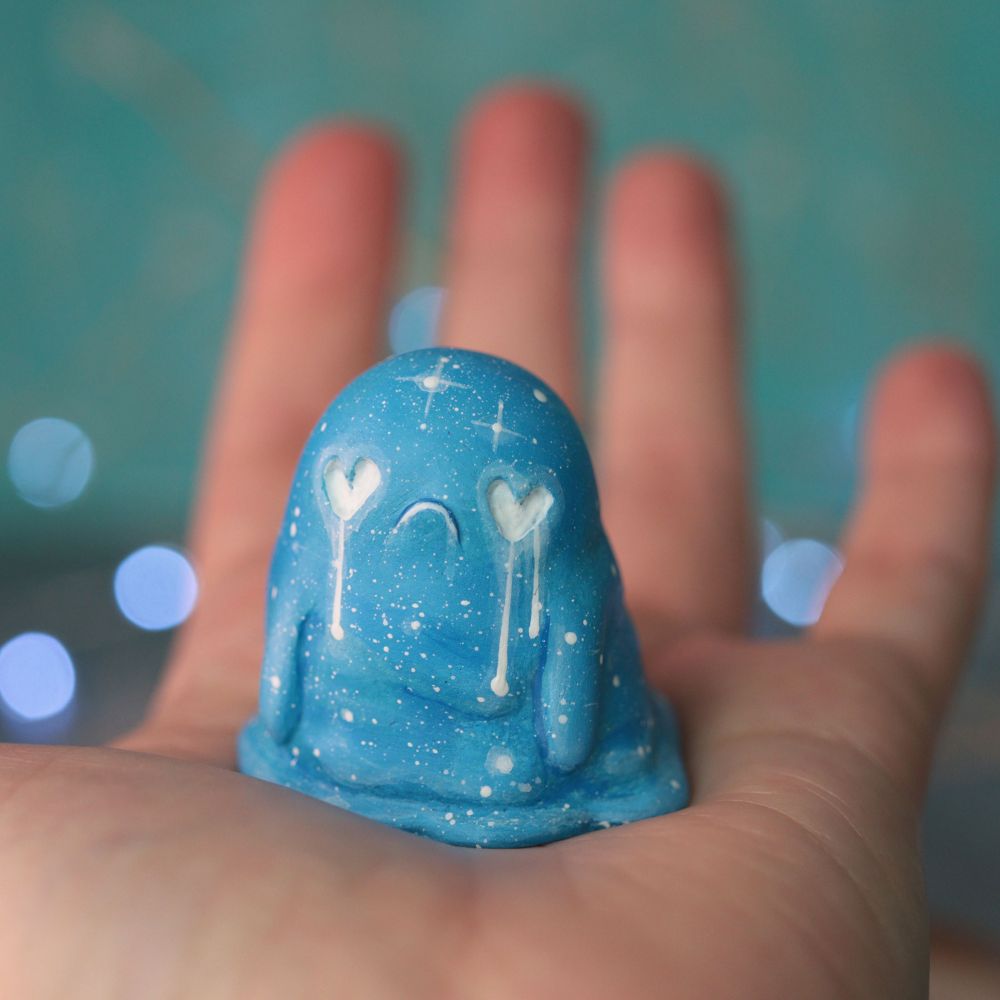 A hand holding a small ghost figurine. The ghost is blue and it has heart shaped eyes with white tears dripping down