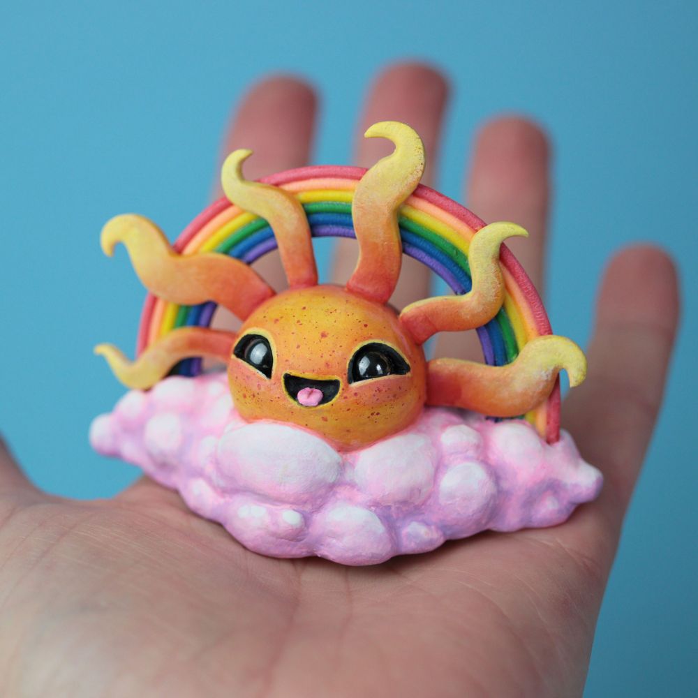 A hand holding a sculpture of a pink cloud with a smiling sun and a rainbow behind its rays. 