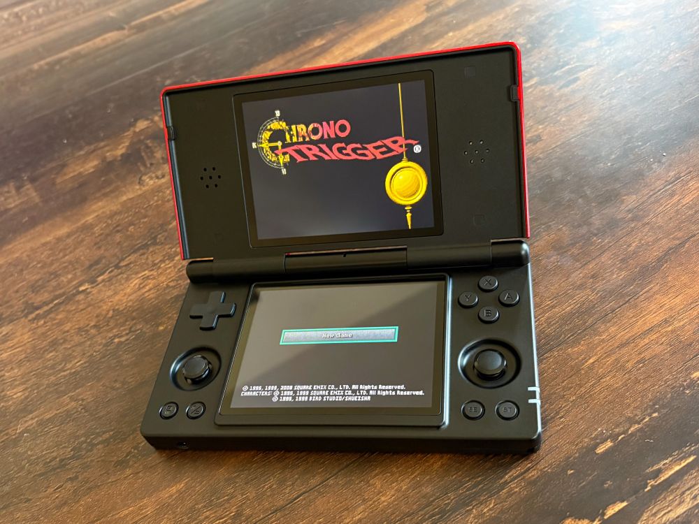 Screenshot of a dual-screened gaming device on a wooden table with the top screen displaying the title for Chrono Trigger as the lower screen is showing the option to start a new game.