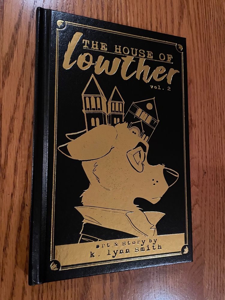 Photo of the second volume of The House of Lowther. It is a handsome hardbound volume with gold foil embossing of the title and an image of the werewolf in a dress shirt in profile looking right with the buildings of the sanitarium sticking out behind his head at odd angles