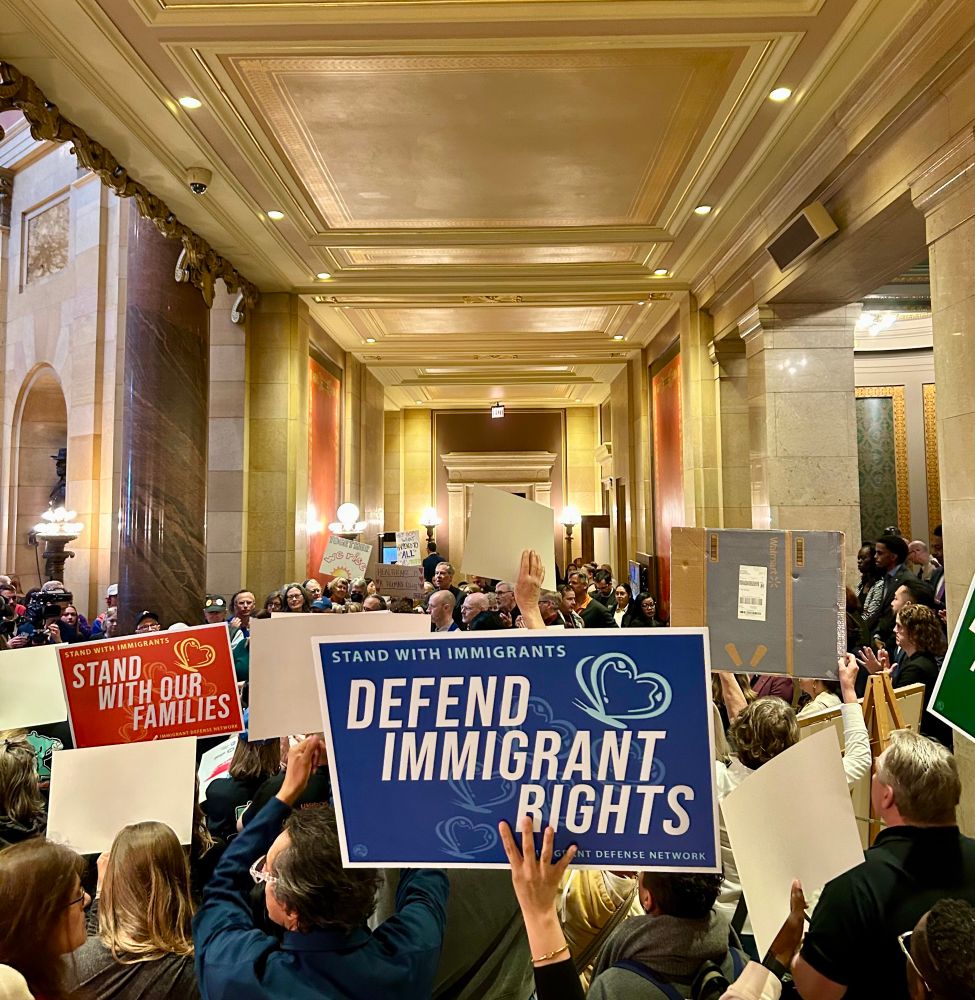 Image of a rally in the Minnesota Capitol, a full hallway of hands and signs. In the foreground is a blue sign reading “Defend Immigrant Rights”