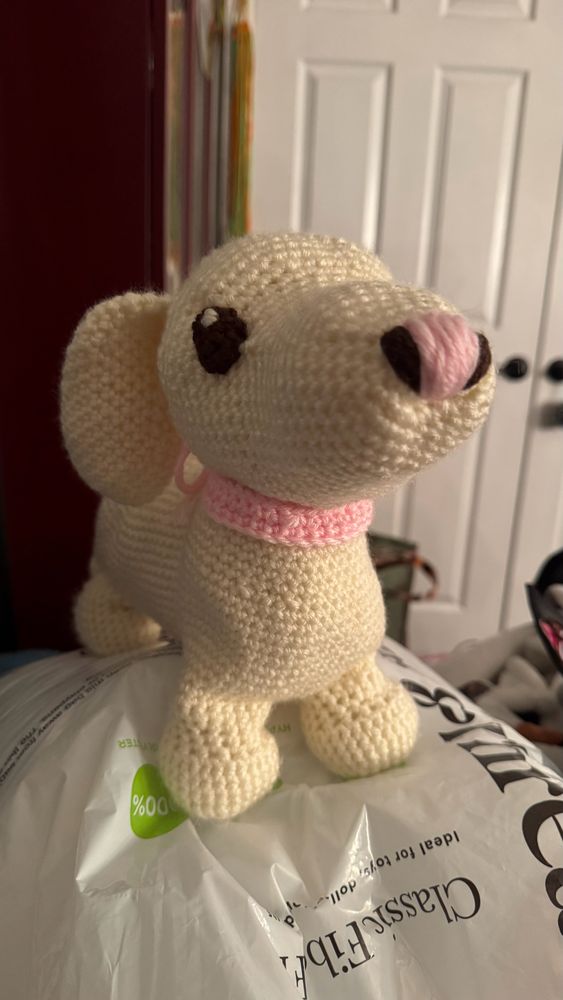 A photo of a white crochet dog with a pink and brown nose, brown eyes and a pink collar. She gazes majestically off into the distance