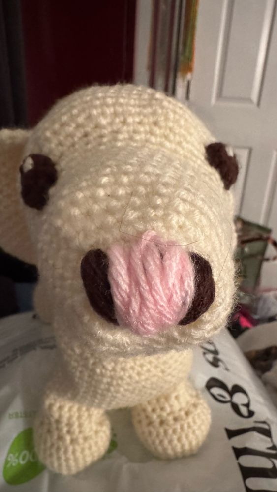 A photo of a white crochet dog with a pink and brown nose, brown eyes and a pink collar. The picture is taken very close to her nose so her snoot looks as long as possible. If you listen closely enough you can hear her one (1) braincell playing pong inside her skull. 