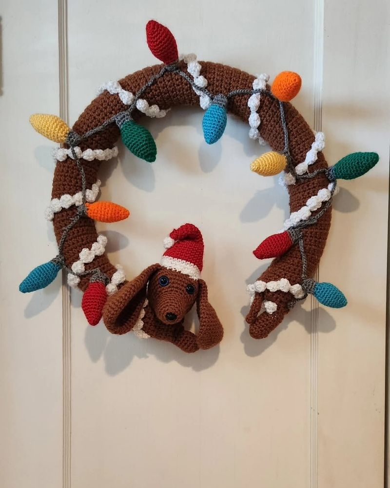 Guirlanda de Natal em formato de cachorro salsicha, onde o corpinho comprido dele forma o círculo da guirlanda e ele tá todo enfeitado com luzes de Natal. na cabeça do cachorro salsicha um gorro de Natal. tudo isso feito de crochê. 