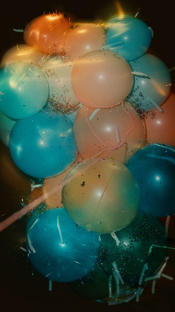 A computer graphics render of a cluster of multicolored balloons floating in black space. Silly string is being shot at the balloons from off screen and glitter is floating in the air around the balloons. The render is meant to look like flash photography from an old point and shoot film camera. 