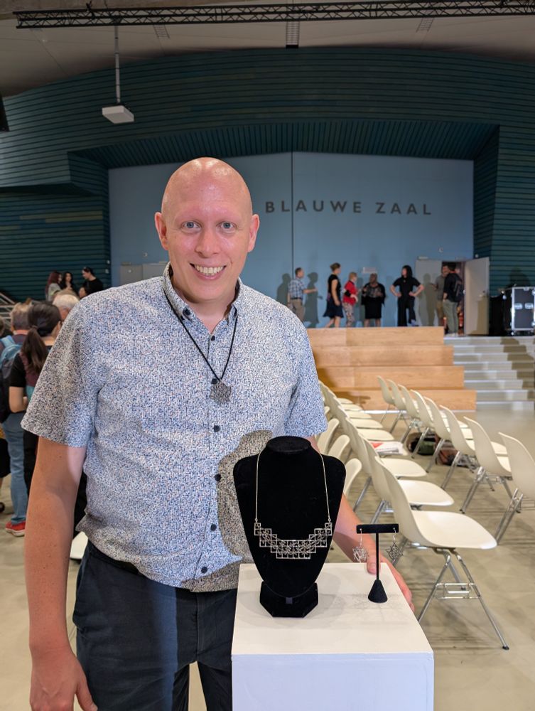 A bald white man standing next to a stand containing the jewelry. 