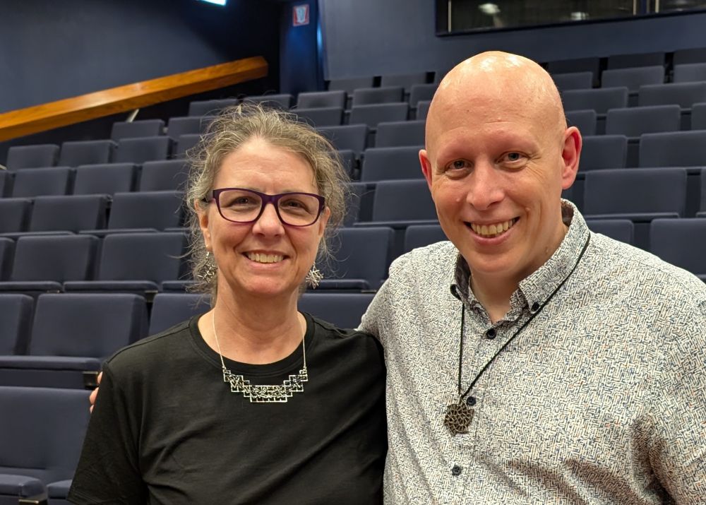 A white woman with grey hair wearing black clothes and the jewelry next to the same bald white man