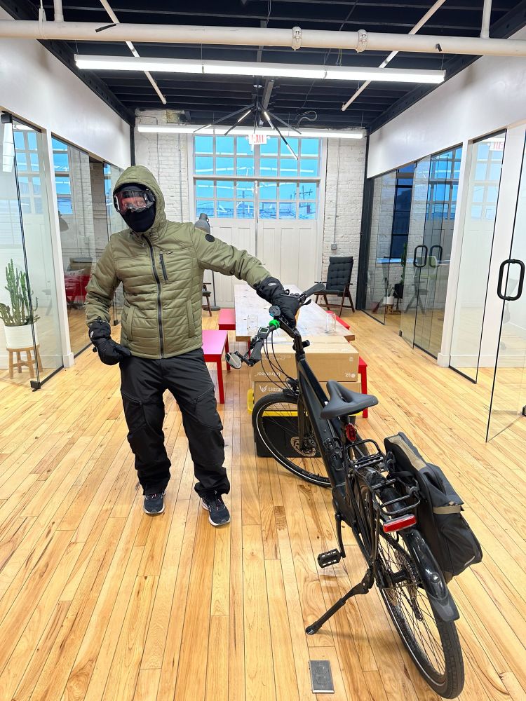Bundled up with snow pants, coat, balaclava, ski goggles, and heavy gloves. Standing next to a bike in an office. 