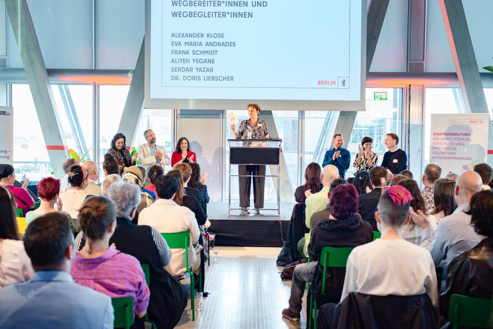 Ein Saal mit den Gästen der der Feier anlässlich des 5-jährigen Jubiläums des Berliner Landesantidiskriminierungsgesetzes. Vorner an der Bühne steht eine Rednerin und hinter ihr an der Leinwand der Text: "Wegbereiter*innen und Wegbegleiter*innen 
Alexander Klose
Eve Maria Andrades
Frank Schmidt 
Aliyeh Yegane
Serdar Yazar
Dr. Doris Liebscher"

Foto: Anke Peters