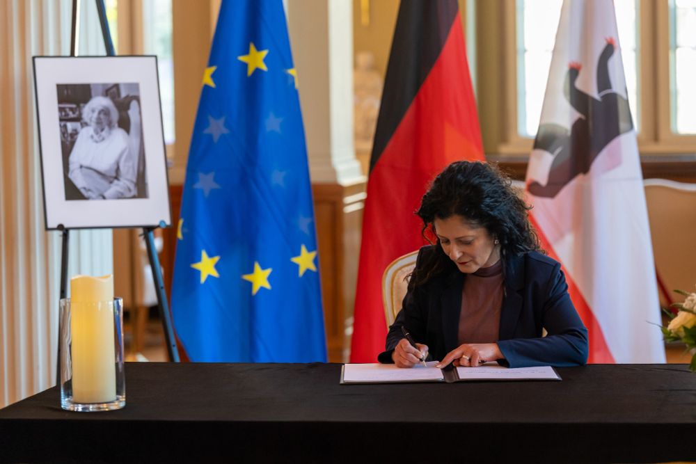 Senatorin für Arbeit, Soziales, Gleichstellung, Integration, Vielfalt und Antidiskriminierung Cansel Kiziltepe sitzt am Tisch im Säulensaal des Roten Rathauses und trägt sich in das Kondolenzbuch ein. Auf dem Tisch liegt eine schwarze Tischdecke und eine große Kerze steht darauf. Hinter dem Tisch steht eine Steffelei mit dem Portrait der verstorbenen Margot Friedländer. Hinter dem Tisch stehen die Flaggen der EU, Deutschlands und Berlins. 

Foto: Landesarchiv Berlin/Grönboldt
