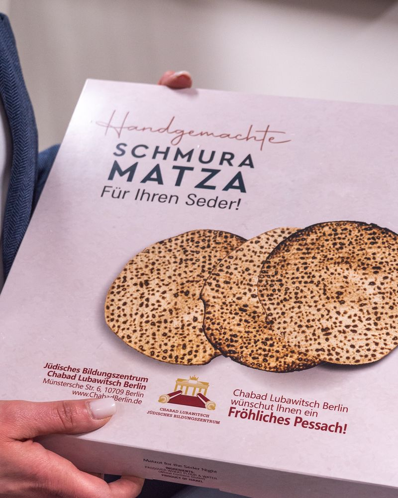 Eine Hand hält einen weißen Karton mit dem bild von drei runden Matze-Broten und dem Text: "Handgemachte Schmura Matza für Ihren Seder
Chabad Lubawitch Berlin wünscht Ihnen ein fröhliches Pessach".
