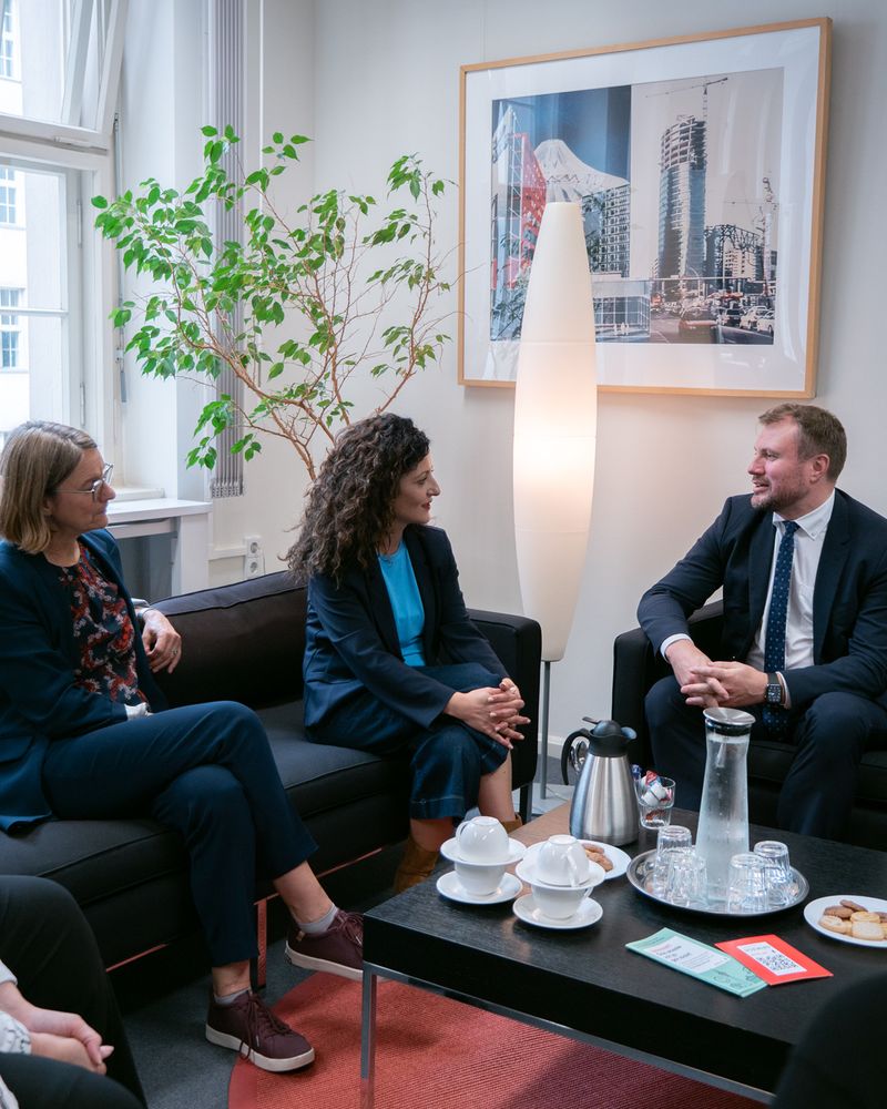 (v.l.n.r.) Katja Karger, Cansel Kiziltepe und Daniel Keller sitzen auf schwarzen Sofas im Amtszimmer der Senatorin zu einem Gespräch zusammen. Auf dem Tisch vor ihnen stehen Kaffeetassen, Wasserkrug und Gebäck. Im Hintergrund: ein großes Fenster, eine Zimmerpflanze und ein gerahmtes Foto mit Stadtansichten.