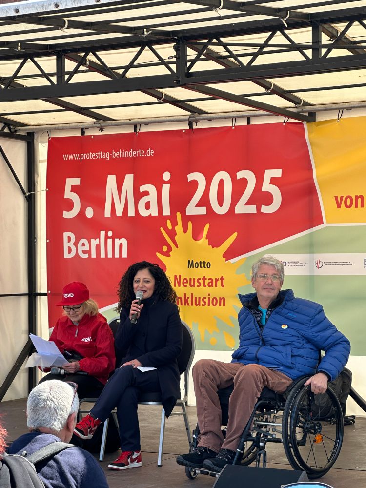 Senatorin Cansel Kiziltepe sitzt auf einer Bühne, links und rechts neben ihr sitzt jeweils eine Person. Die Person auf der rechten Seite sitzt im Rollstuhl. Auf der Rückwand der Bühne steht www.protesttag-behinderte.de und 5. Mai 2025 Berlin - Motto Neustart Inklusion