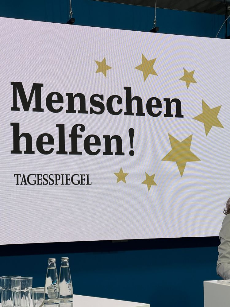 Ein Foto von der weißen Leinwand, an der die große Überschrift "Menschen Helfen!" umrandet von gelben Sternen und darunter "Tagesspiegel" zu sehen sind.