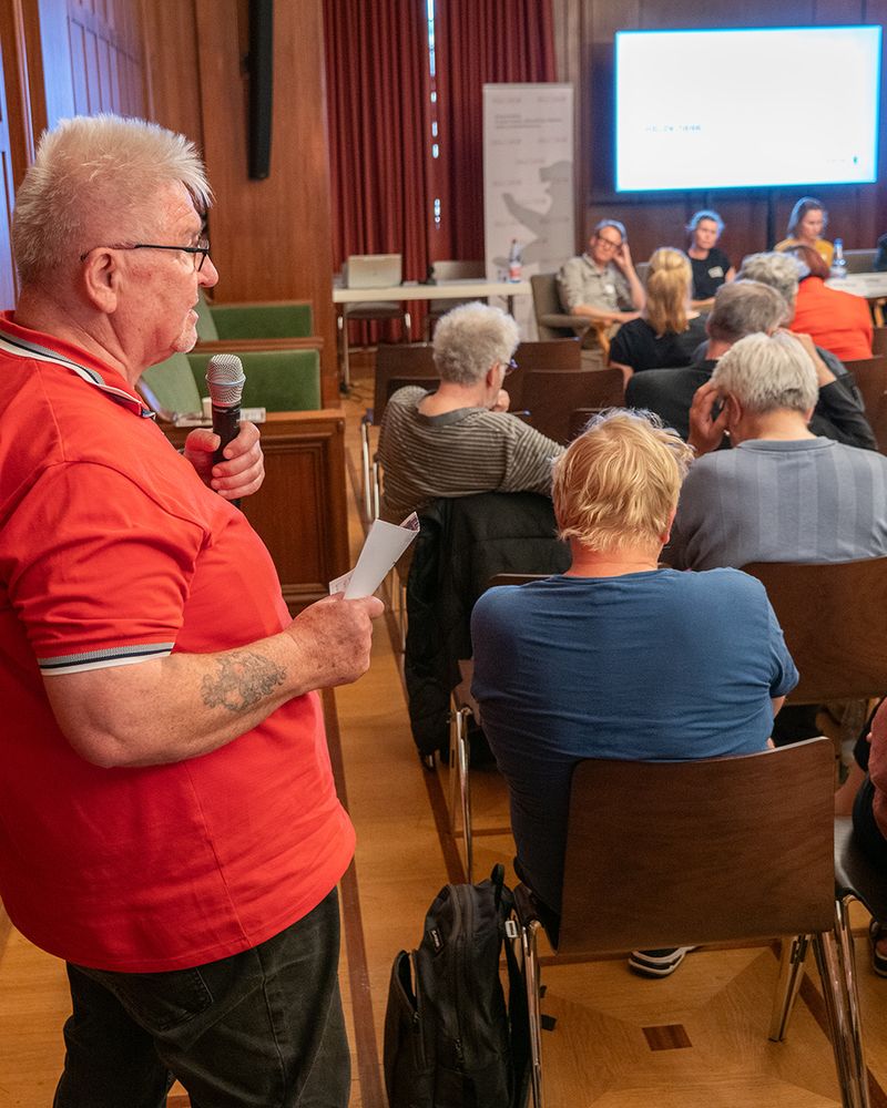 Ein älterer Gast der Veranstaltung hält das Mikrofon und stellt eine Frage. Im Hintergrund sieht man das Publikum der Veranstaltung sowie die Gäste auf dem Panel.
