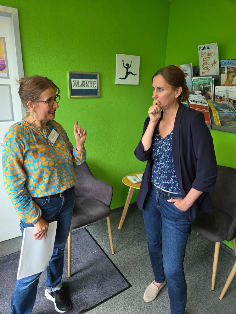 Nina-Sybil Klüppel und Micha Klapp stehen im Gespräch nebeneinander vor einer grünen Wand mit Broschüren und Bildern. Frau Klüppel spricht gestikulierend, während die Staatssekretärin Klapp nachdenklich zuhört. Beide wirken engagiert und inhaltlich vertieft.